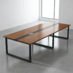 12-seater boardroom table featuring a long rectangular tabletop finished in a warm teak wood grain veneer, styled with a contrasting black running center stripe that houses two silver flip-top cable management grommets, all supported by a heavy-duty industrial matte black metal sled base.
