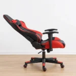 Ergonomic red gaming chair shown from front and side-profile reclined angles, featuring a high-back racing-style silhouette upholstered in black faux leather with vibrant red side panels, equipped with a removable neck pillow, an adjustable lumbar support cushion, T-shaped height-adjustable armrests, a reclining mechanism lever, and a matching five-star nylon swivel base on color-coordinated red-rimmed dual-wheel casters.