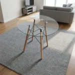 Round eames table featuring a transparent, circular tempered glass tabletop, supported by a classic mid-century modern base composed of four dowel legs in natural light wood, reinforced with an architectural black metal Eiffel wire cross-brace structure and metallic glass-mounting brackets.