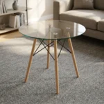 Round eames table featuring a transparent, circular tempered glass tabletop, supported by a classic mid-century modern base composed of four dowel legs in natural light wood, reinforced with an architectural black metal Eiffel wire cross-brace structure and metallic glass-mounting brackets.