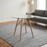 Round eames table featuring a transparent, circular tempered glass tabletop, supported by a classic mid-century modern base composed of four dowel legs in natural light wood, reinforced with an architectural black metal Eiffel wire cross-brace structure and metallic glass-mounting brackets.