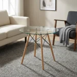 Round eames table featuring a transparent, circular tempered glass tabletop, supported by a classic mid-century modern base composed of four dowel legs in natural light wood, reinforced with an architectural black metal Eiffel wire cross-brace structure and metallic glass-mounting brackets.