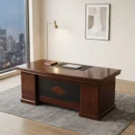 1.8m L-shaped executive office desk suite in a polished cherry wood finish, featuring a thick double-layered main desktop with a central black leather writing pad insert and dual circular cable grommets, supported by a decorative front modesty panel with a black segmented pattern and a central diamond-shaped wood inlay. The workstation is configured with a mobile under-desk two-drawer pedestal cabinet on casters and an attached right-side return credenza with integrated storage, paired with a matching tall cherry wood bookcase with four glass-front upper doors in the background.