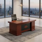 1.8m L-shaped executive office desk suite in a polished cherry wood finish, featuring a thick double-layered main desktop with a central black leather writing pad insert and dual circular cable grommets, supported by a decorative front modesty panel with a black segmented pattern and a central diamond-shaped wood inlay. The workstation is configured with a mobile under-desk two-drawer pedestal cabinet on casters and an attached right-side return credenza with integrated storage, paired with a matching tall cherry wood bookcase with four glass-front upper doors in the background.