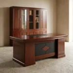 1.8m L-shaped executive office desk suite in a polished cherry wood finish, featuring a thick double-layered main desktop with a central black leather writing pad insert and dual circular cable grommets, supported by a decorative front modesty panel with a black segmented pattern and a central diamond-shaped wood inlay. The workstation is configured with a mobile under-desk two-drawer pedestal cabinet on casters and an attached right-side return credenza with integrated storage, paired with a matching tall cherry wood bookcase with four glass-front upper doors in the background.