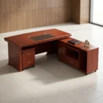 1.8m L-shaped executive office desk suite in a polished cherry wood finish, featuring a thick double-layered main desktop with a central black leather writing pad insert and dual circular cable grommets, supported by a decorative front modesty panel with a black segmented pattern and a central diamond-shaped wood inlay. The workstation is configured with a mobile under-desk two-drawer pedestal cabinet on casters and an attached right-side return credenza with integrated storage, paired with a matching tall cherry wood bookcase with four glass-front upper doors in the background.