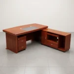 1.8m L-shaped executive office desk suite in a polished cherry wood finish, featuring a thick double-layered main desktop with a central black leather writing pad insert and dual circular cable grommets, supported by a decorative front modesty panel with a black segmented pattern and a central diamond-shaped wood inlay. The workstation is configured with a mobile under-desk two-drawer pedestal cabinet on casters and an attached right-side return credenza with integrated storage, paired with a matching tall cherry wood bookcase with four glass-front upper doors in the background.