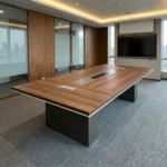 2.4m conference meeting table featuring a natural wood-grain tabletop with white edge trim, a central black media connectivity panel, and dual charcoal gray block pedestal legs.
