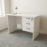 1.0m office study desk featuring a clean white finish, a circular black cable grommet on the tabletop, a matching modesty panel, and an integrated three-drawer pedestal with black curved handles and a lockable top drawer.