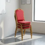Red stackable banquet chair featuring a crown-back backrest with an integrated top carry handle, upholstered in vibrant red fabric with a subtle gold diamond lattice pattern, paired with a thick padded seat cushion and supported by a durable gold-finished metallic frame with protective bumper guards on the legs.