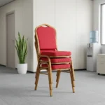 Red stackable banquet chair featuring a crown-back backrest with an integrated top carry handle, upholstered in vibrant red fabric with a subtle gold diamond lattice pattern, paired with a thick padded seat cushion and supported by a durable gold-finished metallic frame with protective bumper guards on the legs.