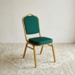 Green stackable banquet chair featuring a crown-top backrest with a cut-out handle and a thick square seat cushion, both upholstered in dark green fabric with a subtle gold dot or diamond pattern, shown in a vertical stack of three supported by a sturdy gold-finished metallic frame with white stacking bumpers.