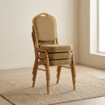 Stackable brown conference chair featuring a crown-top backrest with a cut-out handle and a thick square seat cushion, both upholstered in a textured light-brown or beige patterned fabric, shown in a vertical stack of three supported by a sturdy gold-finished metallic frame with integrated stacking bumpers.