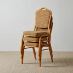 Stackable brown conference chair featuring a crown-top backrest with a cut-out handle and a thick square seat cushion, both upholstered in a textured light-brown or beige patterned fabric, shown in a vertical stack of three supported by a sturdy gold-finished metallic frame with integrated stacking bumpers.
