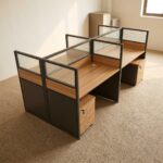 4-way modular workstation featuring four light oak-finish desks arranged in a 2x2 cluster, separated by semi-private partition screens with dark gray lower panels, light oak accent trim, and frosted glass upper sections, complete with individual under-desk three-drawer mobile pedestals and integrated desktop cable grommets.