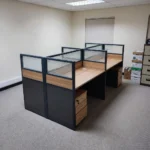 4-way modular workstation featuring four light oak-finish desks arranged in a 2x2 cluster, separated by semi-private partition screens with dark gray lower panels, light oak accent trim, and frosted glass upper sections, complete with individual under-desk three-drawer mobile pedestals and integrated desktop cable grommets.