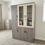 3-door wooden executive cabinet featuring a two-tone finish with taupe wood-grain laminate, upper cream-framed glass display doors revealing internal shelving, lower solid wooden doors, modern dark vertical handles, and key-lock security mechanisms.