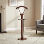 Vertical wooden valet stand featuring a dark mahogany-finished frame, an integrated contoured coat hanger supported by metallic struts, a horizontal trouser bar with wooden end caps, a central vertical stem, and a sturdy round wooden base.