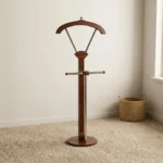 Vertical wooden valet stand featuring a dark mahogany-finished frame, an integrated contoured coat hanger supported by metallic struts, a horizontal trouser bar with wooden end caps, a central vertical stem, and a sturdy round wooden base.