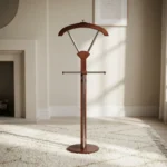 Vertical wooden valet stand featuring a dark mahogany-finished frame, an integrated contoured coat hanger supported by metallic struts, a horizontal trouser bar with wooden end caps, a central vertical stem, and a sturdy round wooden base.