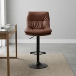 Brown leather adjustable barstool featuring a plush, tufted warm brown leatherette seat and backrest, supported by a matte black pedestal stem, an integrated circular footrest, pneumatic gas-lift height adjustment, and a round matte black base.