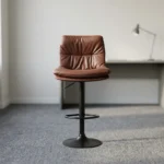 Brown leather adjustable barstool featuring a plush, tufted warm brown leatherette seat and backrest, supported by a matte black pedestal stem, an integrated circular footrest, pneumatic gas-lift height adjustment, and a round matte black base.