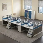 6-way office workstation featuring six light grey desktop surfaces arranged face-to-face, framed partition screens with blue fabric and glass top panels, side modesty panels, and six under-desk white three-drawer mobile pedestals.