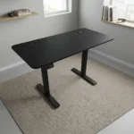 Height adjustable electric table featuring a sleek matte black desktop with dual cable management grommets, an under-desk digital control keypad, and heavy-duty black motorized telescoping legs supported by T-style flat feet.