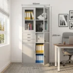 White office cabinet with safe featuring a clean white finish, top display section with two glass-framed lockable doors, middle section equipped with three small pull-out drawers alongside a lockable safe compartment, and a bottom storage section with two solid doors with vertical handles and key locks.