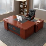 Executive 2m office table featuring a rich mahogany wood grain finish, an inset black leatherette writing pad with integrated wire management grommets, a patterned front modesty panel with leatherette accent panels, sturdy pedestal end legs on dark bases, and an attached side return cabinet.