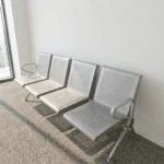 Heavy duty 4-seater waiting bench featuring a durable metallic silver-grey steel construction, four individual perforated metal seats and backrests, curved side armrests, a heavy-duty central support beam, and angled steel legs.