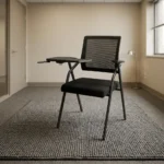 Foldable chair with writing pad featuring a breathable black mesh backrest, a cushioned dark grey fabric seat, integrated armrests, a built-in foldaway writing tablet with a cup holder, and a space-saving foldable black tubular steel frame.