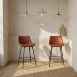 Leather kitchen bar stool featuring warm cognac brown faux leather upholstery with vertical channel tufting, a low-profile ergonomic backrest, and slim matte black metal legs with integrated footrest rungs.
