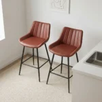 Leather kitchen bar stool featuring warm cognac brown faux leather upholstery with vertical channel tufting, a low-profile ergonomic backrest, and slim matte black metal legs with integrated footrest rungs.