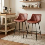 Leather kitchen bar stool featuring warm cognac brown faux leather upholstery with vertical channel tufting, a low-profile ergonomic backrest, and slim matte black metal legs with integrated footrest rungs.