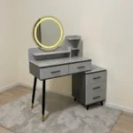 Modern dressing table with LED featuring a circular touch-sensor illuminated vanity mirror, a grey faux-marble tabletop, fluted grey drawer fronts with black handles, built-in display shelves, dark tapered legs with gold accents, and a side three-drawer storage chest.