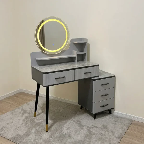 Modern dressing table with LED featuring a circular touch-sensor illuminated vanity mirror, a grey faux-marble tabletop, fluted grey drawer fronts with black handles, built-in display shelves, dark tapered legs with gold accents, and a side three-drawer storage chest.