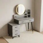 Modern dressing table with LED featuring a circular touch-sensor illuminated vanity mirror, a grey faux-marble tabletop, fluted grey drawer fronts with black handles, built-in display shelves, dark tapered legs with gold accents, and a side three-drawer storage chest.