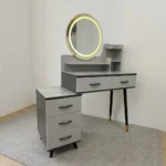 Modern dressing table with LED featuring a circular touch-sensor illuminated vanity mirror, a grey faux-marble tabletop, fluted grey drawer fronts with black handles, built-in display shelves, dark tapered legs with gold accents, and a side three-drawer storage chest.