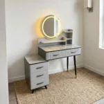 Modern dressing table with LED featuring a circular touch-sensor illuminated vanity mirror, a grey faux-marble tabletop, fluted grey drawer fronts with black handles, built-in display shelves, dark tapered legs with gold accents, and a side three-drawer storage chest.