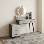 Modern dressing table with LED featuring a circular touch-sensor illuminated vanity mirror, a grey faux-marble tabletop, fluted grey drawer fronts with black handles, built-in display shelves, dark tapered legs with gold accents, and a side three-drawer storage chest.