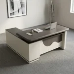 Spacious 1.6m office table featuring a two-tone grey wood-grain and cream laminate surface with rounded edges, a rectangular cable management outlet, an L-shaped side return credenza with open shelving and lockable drawers, and a decorative front modesty panel with a distinctive wave-pattern accent.