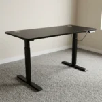 Height adjustable standing desk featuring a sleek matte black rectangular desktop, dual circular cable management grommets, an under-desk digital control keypad, and a motorized black T-frame steel base with telescoping legs and wide stabilizing feet.