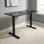 Height adjustable standing desk featuring a sleek matte black rectangular desktop, dual circular cable management grommets, an under-desk digital control keypad, and a motorized black T-frame steel base with telescoping legs and wide stabilizing feet.