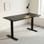 Height adjustable standing desk featuring a sleek matte black rectangular desktop, dual circular cable management grommets, an under-desk digital control keypad, and a motorized black T-frame steel base with telescoping legs and wide stabilizing feet.