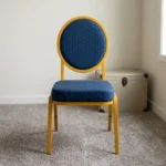 Armless blue conference chair featuring navy blue fabric upholstery with white dot pattern, a oval medallion backrest, a cushioned seat, and a stackable gold-finished metal frame with straight legs and floor glides.