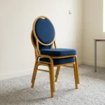 Armless blue conference chair featuring navy blue fabric upholstery with white dot pattern, a oval medallion backrest, a cushioned seat, and a stackable gold-finished metal frame with straight legs and floor glides.