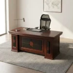 Mahogany 1.6m executive desk featuring a rich wood veneer finish with bevelled edge detailing, an inset black leatherette writing pad, dual desktop cable management grommets, an ornate decorative front modesty panel with black leatherette inlay and circular wood accents, and thick pedestal leg supports.