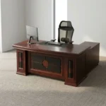 Mahogany 1.6m executive desk featuring a rich wood veneer finish with bevelled edge detailing, an inset black leatherette writing pad, dual desktop cable management grommets, an ornate decorative front modesty panel with black leatherette inlay and circular wood accents, and thick pedestal leg supports.