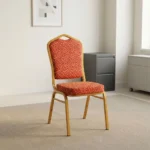 Fabric stackable conference chair featuring patterned red and gold patterned fabric upholstery, high-density padded seat and crown-back cushion, an armless space-saving stackable frame, an integrated top hand-hold, and a gold powder-coated square steel tube frame with bumper guards and floor glides.
